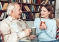 Senior man and young woman at home library sitting on sofa holding cups of hot coffee talking smiling happy